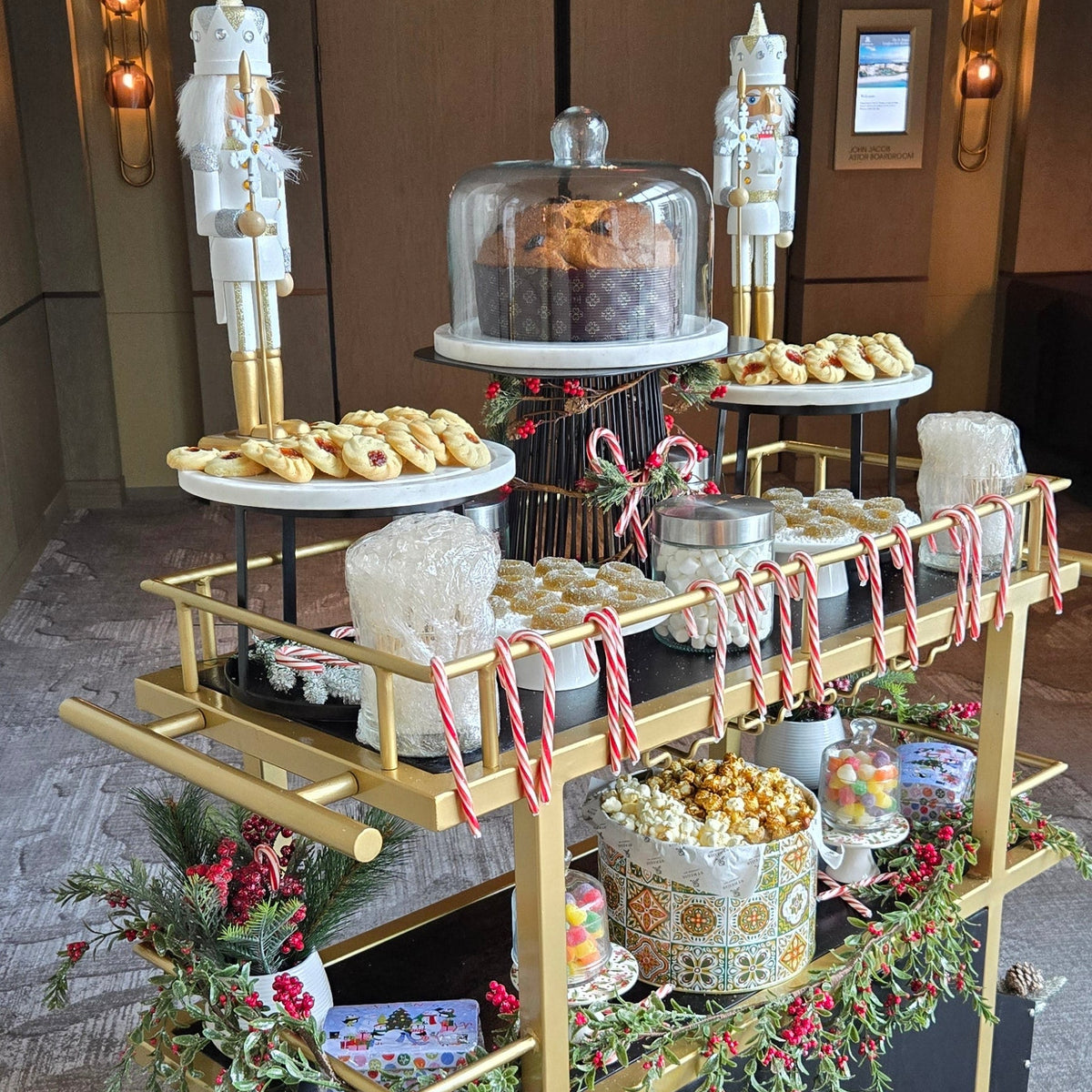 A holiday-themed dessert display cart filled with pastries, candy canes, popcorn, and a covered cake, set indoors with nutcracker decorations and festive garlands.